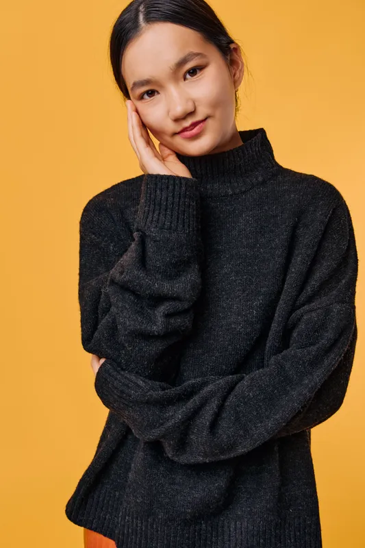 Young woman with hand near face wearing black knitted sweater standing against yellow background. Casual style, smile, long dark hair, portrait, beauty and confidence in studio shot.