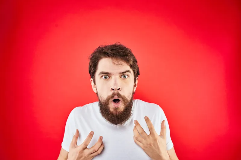 Эмоциональный бородатый мужчина в белой футболке жестикулирует руками, выражая агрессию и недовольство на красном фоне