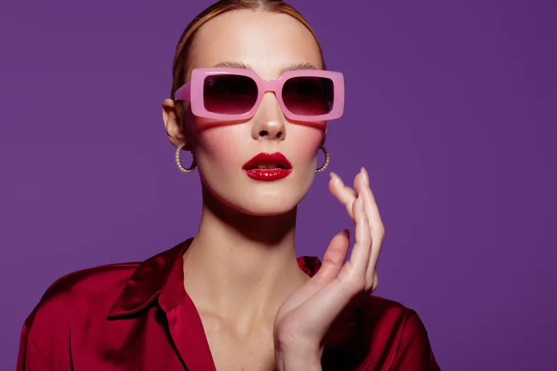 Stylish woman with pink sunglasses and red lipstick poses against purple background. Fashion portrait with glossy makeup and elegant red satin blouse. Concept of glamour and style.