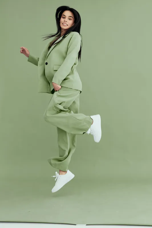 young woman jumping in green suit with white sneakers, smiling, energetic and joyful, isolated on green background, fashion and lifestyle concept