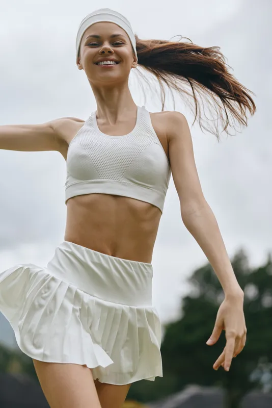 Smiling young woman in workout attire dancing outdoors, showcasing fitness and joy with a dynamic background in soft green tones