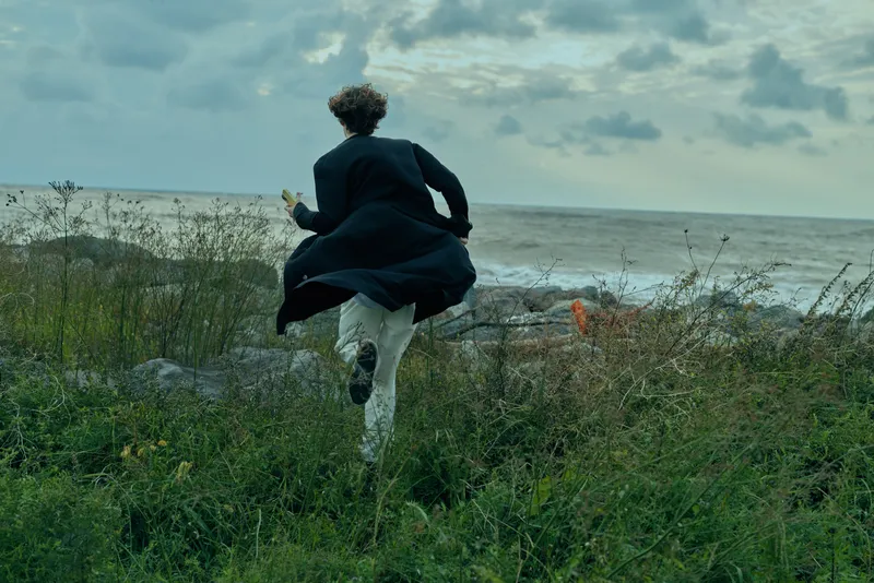Runner in a dark coat dashes across coastal dunes toward the sea, wind whipping around, capturing movement, nature, and solitary escape beside the shoreline