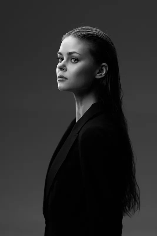 Black and white portrait of serious young woman with long hair wearing dark jacket on gray background.