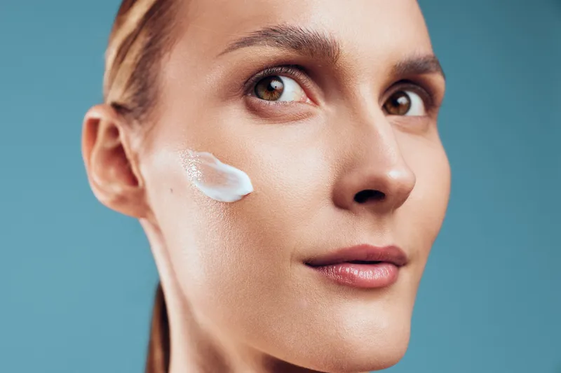 Closeup of woman applying moisturizing cream on cheek, clear skin, natural beauty, soft light, smooth texture, skincare routine, healthy complexion, blue background