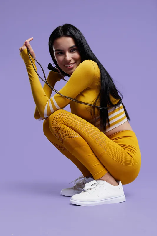 Sport fitness woman in vibrant yellow activewear squatting with jump rope, smiling confidently against purple studio background, promoting healthy lifestyle and exercise.