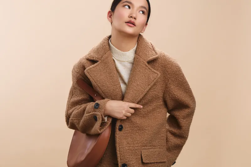 asian woman dressed in a chic coat stands in a modern office setting, smiling as she texts on her phone, creating a bright, positive, and dynamic atmosphere of focused professional communication.