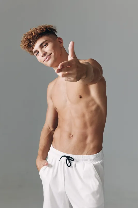 Young man with curly hair shirtless wearing white shorts posing with hand gesture and confident smile on gray background in studio. Male model with athletic body.