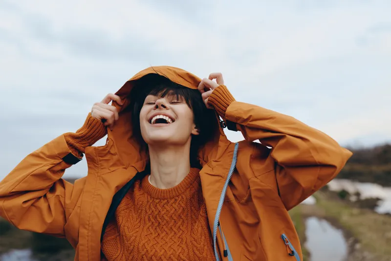 Woman lifestyle outdoors in an orange jacket smiling widely, hands holding a hat, laughing in fresh wind, scenic nature with river and hills under soft daylight, open sky horizon