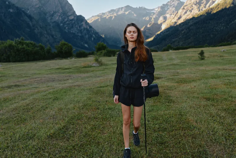 A young woman stands in a wide grassy field with distant mountains. She wears a dark jacket and shorts, a shoulder bag, and looks thoughtful, ready for adventure in a natural landscape.