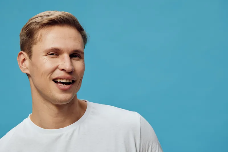 Smiling man with short light brown hair wearing casual white t-shirt against a solid light blue background, studio portrait, cheerful expression, relaxed mood, lifestyle concept, male model
