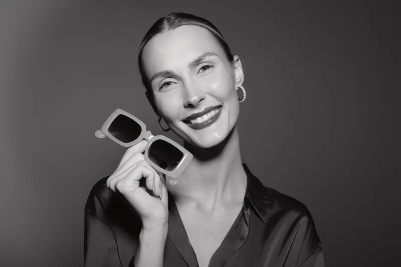 Smiling Caucasian woman with light skin holding sunglasses close to her face, wearing earrings and silky blouse, studio portrait.