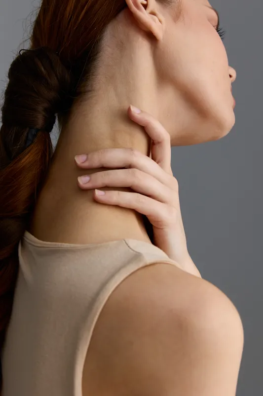 Close-up of a woman’s neck and shoulder with a gentle hand touch, showing smooth skin texture and soft lighting on a neutral gray background.