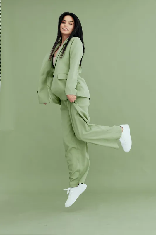 Young woman jumping joyfully wearing a green suit with white sneakers, showcasing happiness and energy against a seamless matching green background in a studio setting.