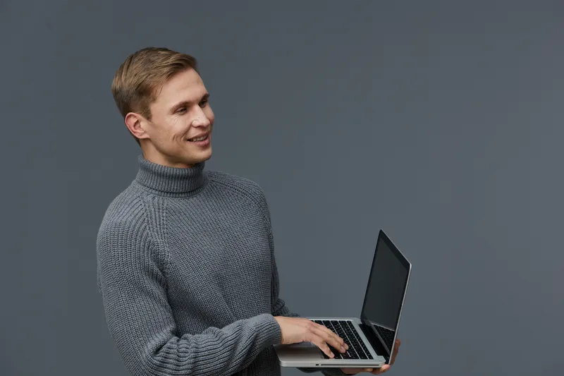Smiling young man with blonde hair wearing a gray turtleneck sweater holding a laptop isolated on plain gray background. Studio portrait of cheerful professional male. People lifestyle concept.