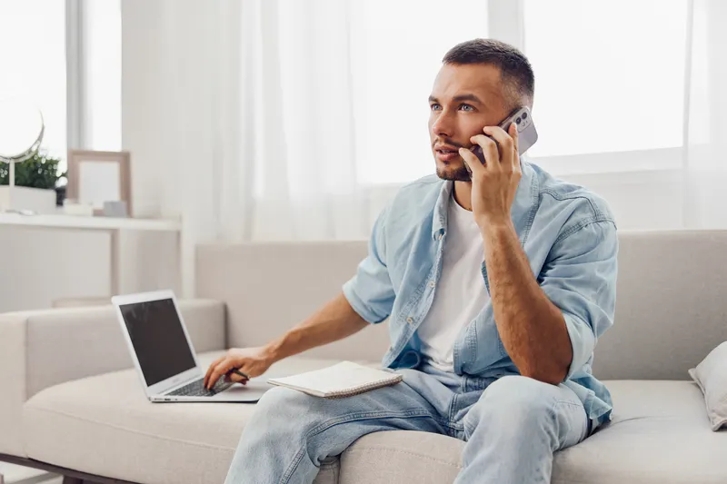 Thoughtful man talking on the phone while working on a laptop, dressed casually in a cozy, modern living space filled with natural light and soft colors.