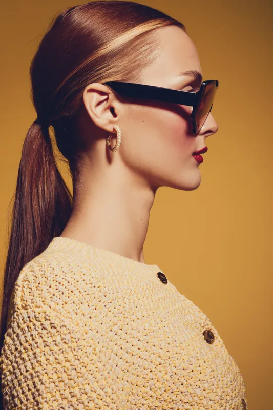 Profile of a stylish woman with long hair tied back wearing large sunglasses and gold hoop earrings. She wears a textured cream sweater against a warm yellow background. Fashion concept.