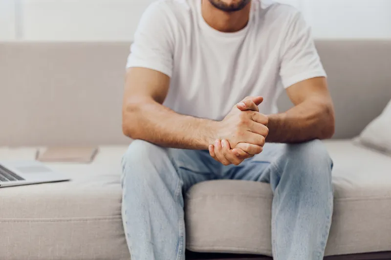 Relaxed man in casual clothing sitting on a cozy sofa with hands clasped, exuding calmness and confidence in a modern living space. Bright and inviting atmosphere.