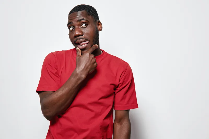 African American man with dark skin wearing red t shirt looking thoughtful with hand on chin isolated on white background. Portrait, expression and casual style.