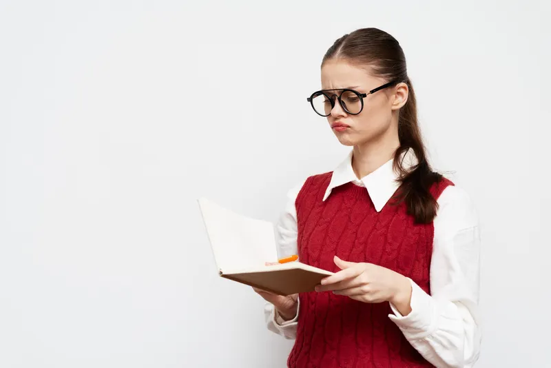 Девушка в очках выглядит озадаченной, читая книгу; на ней красный свитер поверх белой рубашки