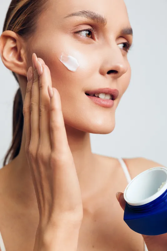 Young woman with clear skin applies white face cream on her cheek while holding a blue jar, expressing gentle care and focus in a bright studio setting with soft lighting.