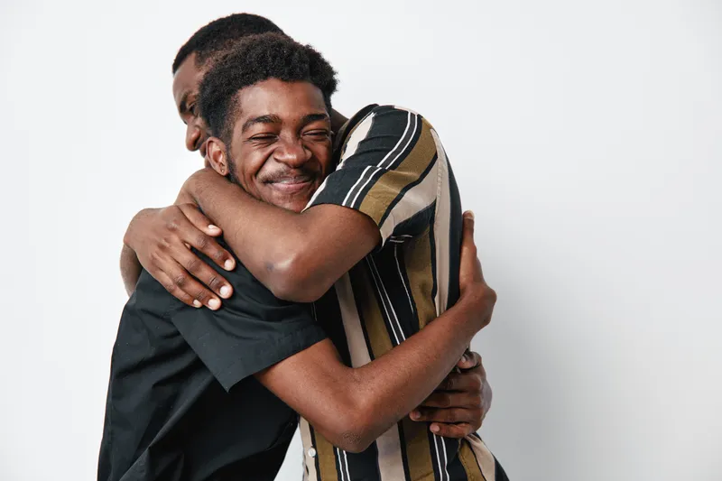 Two young black men hugging each other with smiles on white background. Friendship, affection, and happiness between african american males in casual clothing with close embrace.