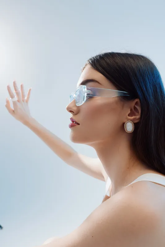 Profile of a stylish woman wearing transparent glasses and pearl earrings. She extends her arm forward against a soft light background. Concept of beauty, fashion, and elegance.