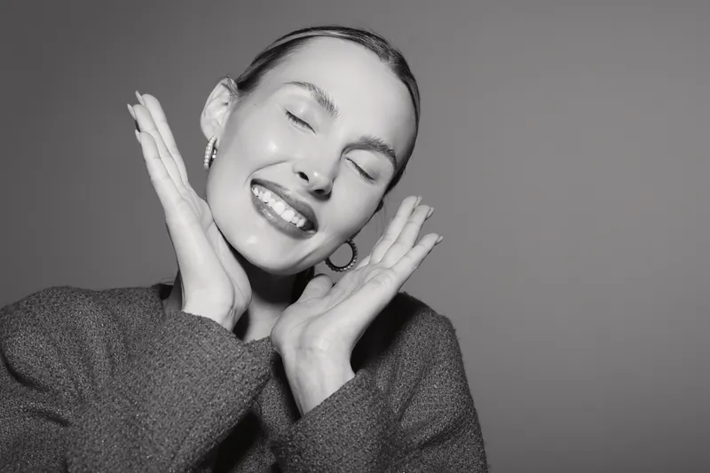 Happy young white woman with light skin smiling with closed eyes and hands near face in studio portrait.