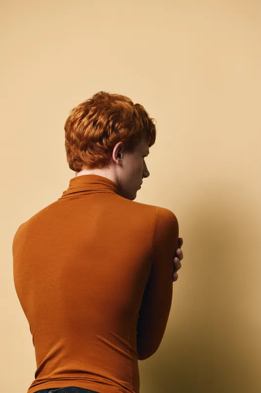 Red hair man in brown turtleneck sweater standing turned back with folded arms on beige background. Portrait from rear with short hairstyle and casual wear for fashion concept.