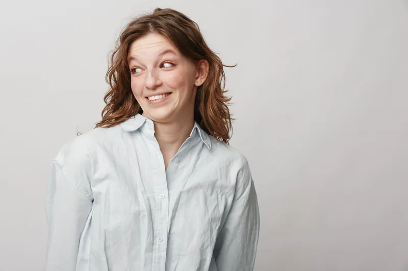 Smiling young woman with curly hair wearing white shirt looks sideways with playful expression on light background. Concept of happiness, casual style, youth and positive emotions.