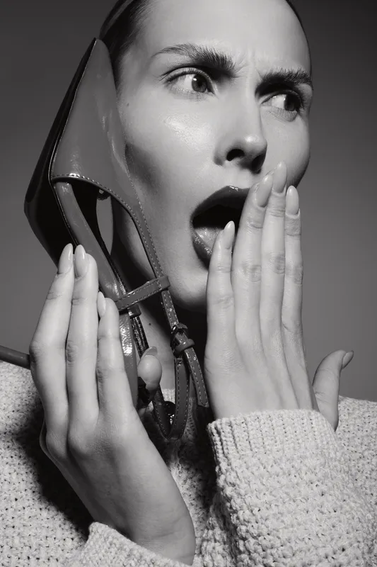 Surprised Caucasian woman with fair skin holding high heel shoe near face in studio black and white fashion concept of emotion and style.