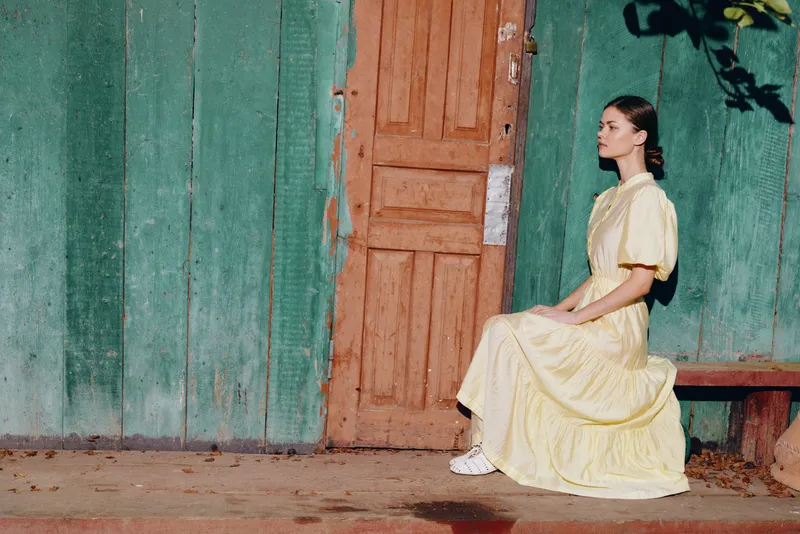 A woman in a light yellow dress sits calmly on a bench beside a rustic door against a green wall outdoors with a thoughtful serene demeanor