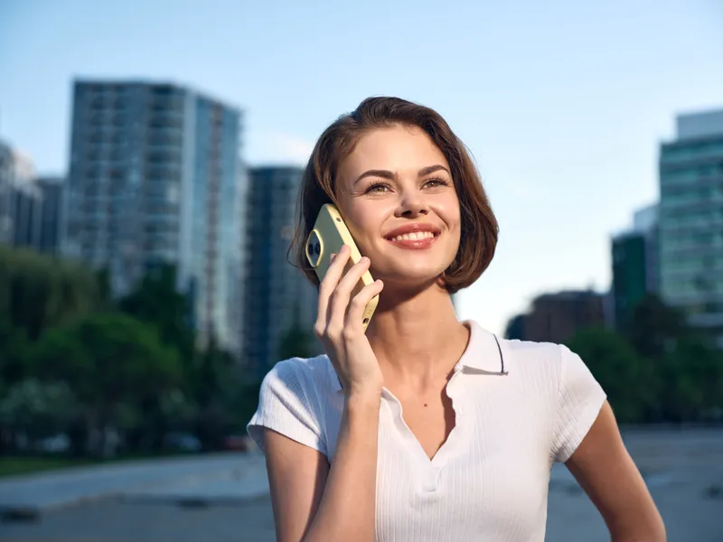 A cheerful young woman talks on a mobile phone outdoors in an urban park setting. She smiles warmly, wearing a light polo shirt, enjoying a sunny day in the city skyline.
