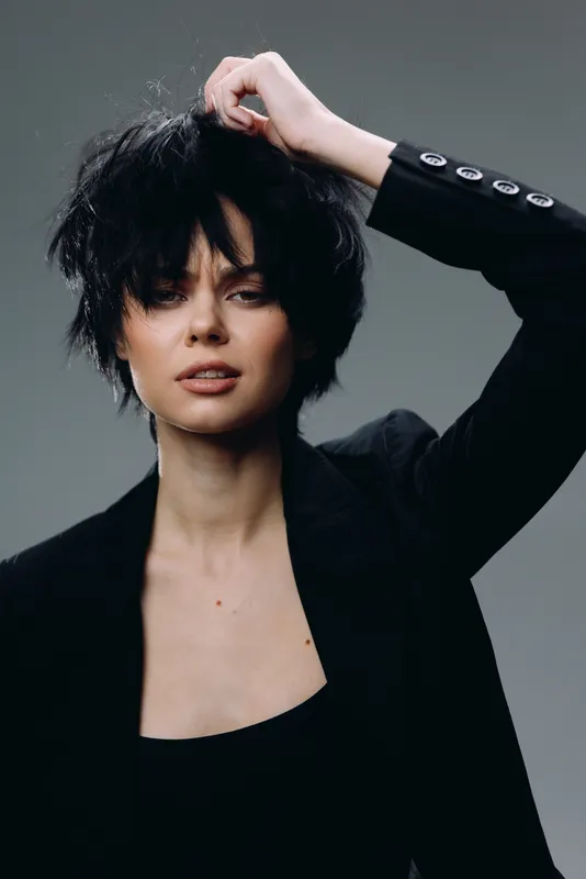 Fashionable woman with short black hair posing confidently in a black blazer against a neutral background, showcasing modern style and attitude.