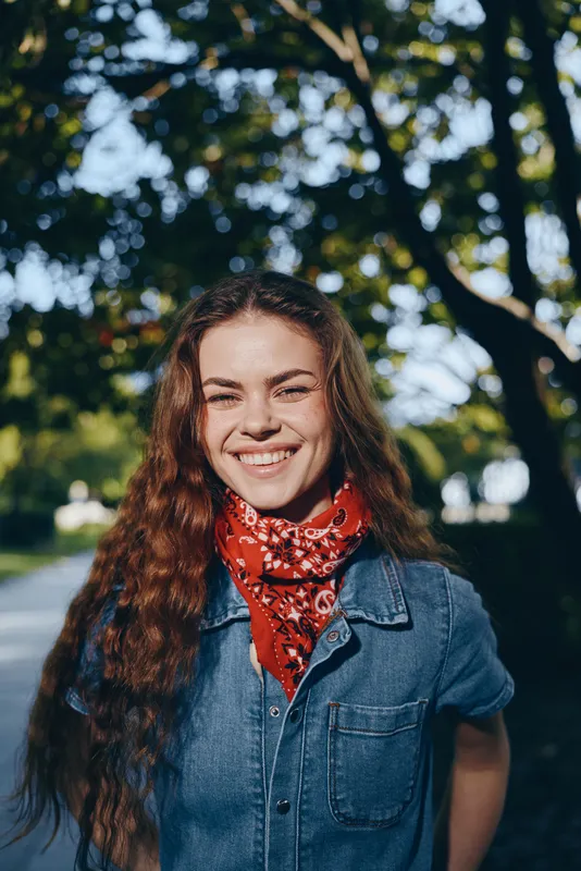 smiling woman with curly hair wearing denim jacket and red scarf outdoors in warm sunlight with blurred green leafy background