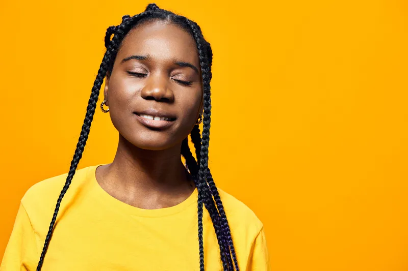 Young African woman with braided hair smiling peacefully in a bright yellow shirt against a vibrant yellow background, conveying joy and positivity