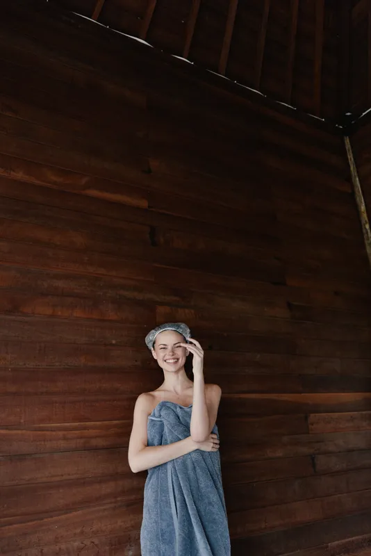 smiling woman wrapped in towel with hair wrap standing against wooden wall, relaxed and fresh indoors, wellness and spa concept
