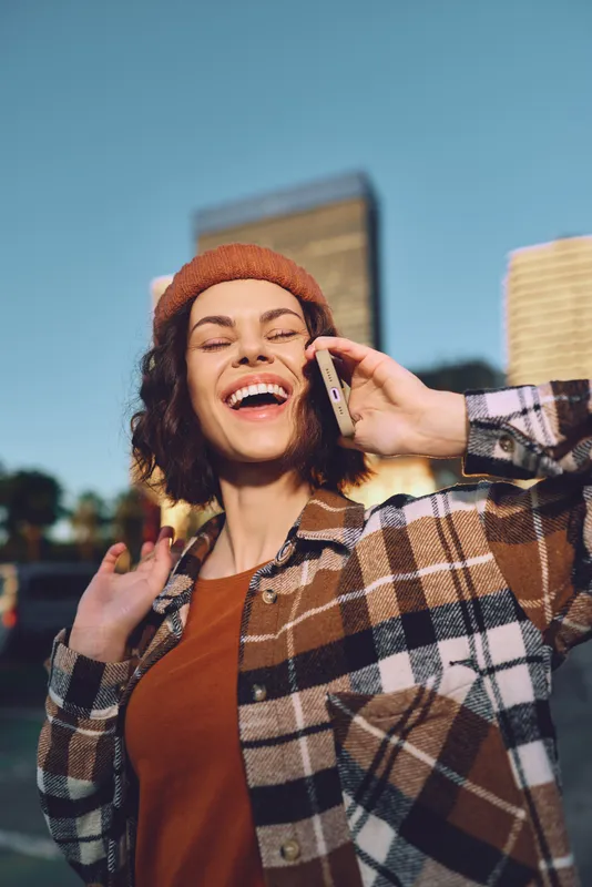 Woman on phone with smile in urban street, candid moment of authenticity and laughter wearing beanie and plaid jacket at golden hour glow, mindful living and emotional storytelling in city life.