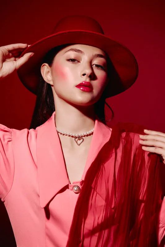 Young woman in pink blazer and red wide-brim hat poses confidently against red background. Dramatic makeup highlights glossy lips and vibrant cheeks. Beauty fashion creative.