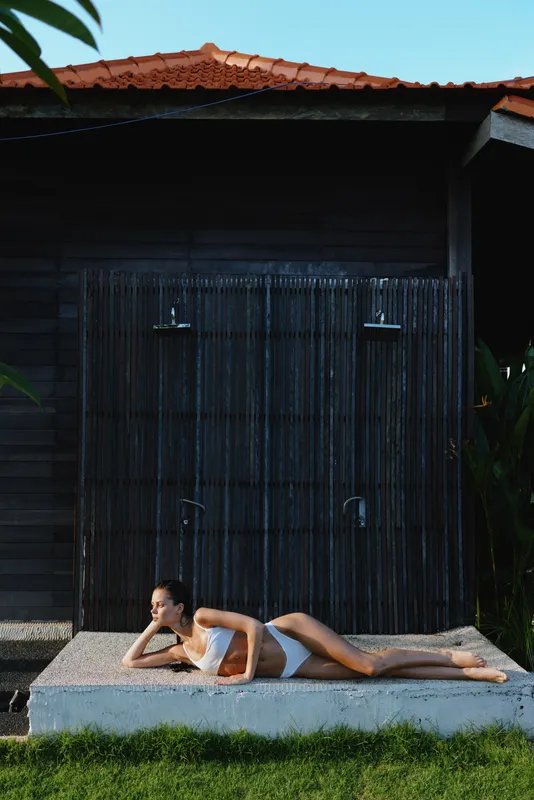 Young woman in white swimsuit lies on concrete platform near dark wooden wall outdoors relaxing in sunny day enjoying summer lifestyle and peaceful moment in tropical environment.