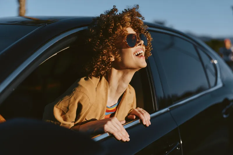 A woman enjoys a sunny car ride with retro film color, leaning out of the window, smiling in sunglasses, wind in her hair, wearing a light jacket. A carefree mood and nostalgic travel vibe.