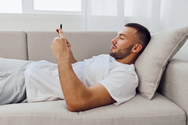 Relaxed man engaging with smartphone at home, reflecting on digital detox and emotional well-being in modern life amid blurred work-life boundaries.