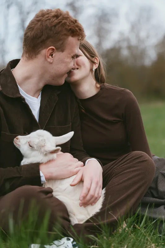 Couple sitting on grass with a friendly goat kid, sharing a tender moment outdoors in bright daylight. Warm natural lighting highlights affection, companionship, and a rustic countryside lifestyle.