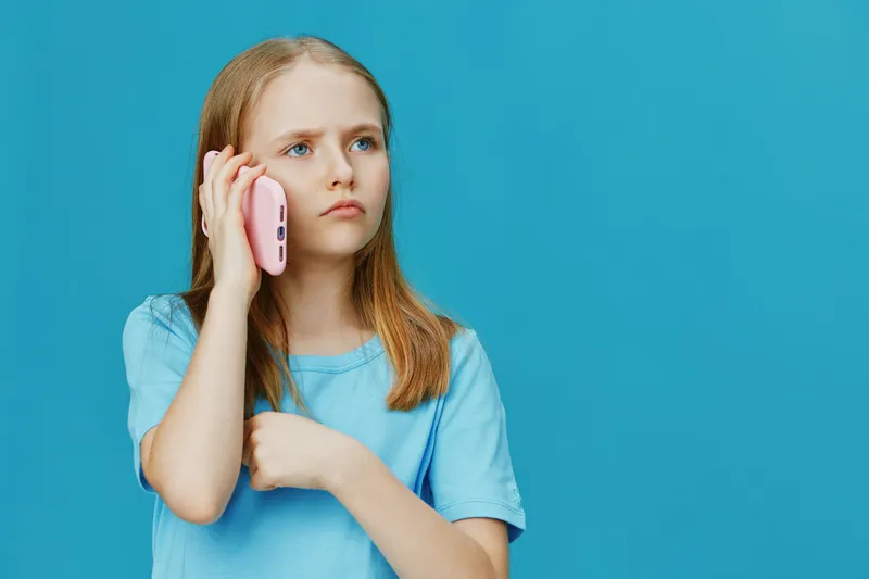 Thoughtful girl talking on pink phone against blue background, expressing curiosity and engagement in conversation, wearing a casual blue shirt