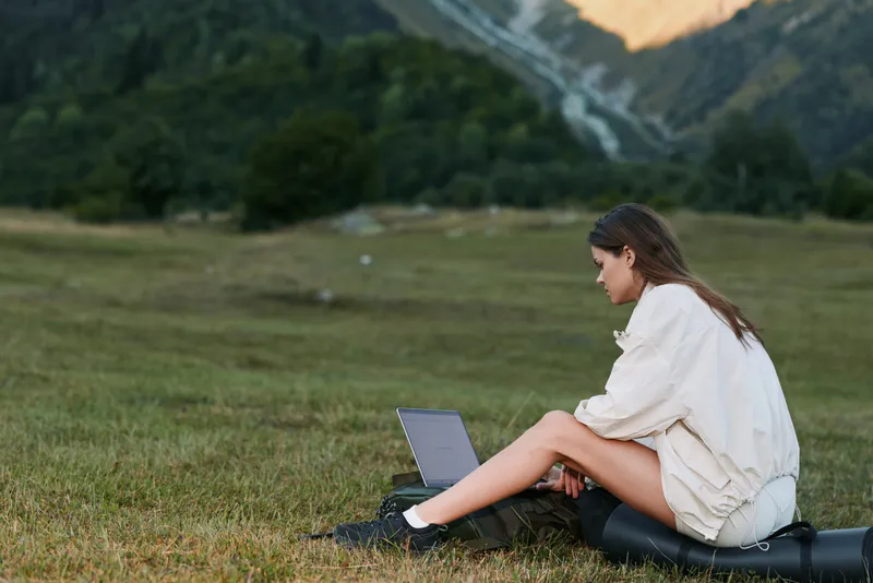 A thoughtful woman sits on a grassy field with a laptop, working outdoors. She wears a light coat, focused on the screen, blending nature with modern digital activity in a calm and productive scene.