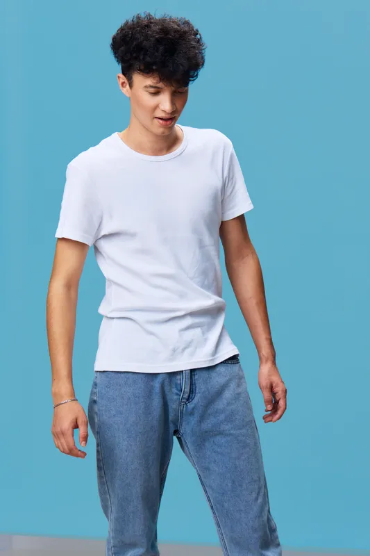 young man wearing casual white t-shirt and blue jeans stands against blue background looking down thoughtfully with curly dark hair and relaxed posture