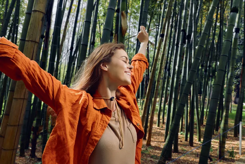 Woman bamboo forest joy arms nature portrait freedom smiling with outstretched arms in orange jacket among tall green bamboo stalks, peaceful outdoor relaxation and mindful breathing.