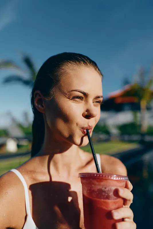 Woman enjoys a refreshing smoothie outdoors on a sunny summer day, sipping through a straw, relaxed lifestyle scene by a pool, casual drinking moment showcasing wellness.