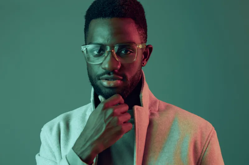 Stylish young African man in glasses posing thoughtfully against a vibrant teal background, showcasing modern fashion and confidence