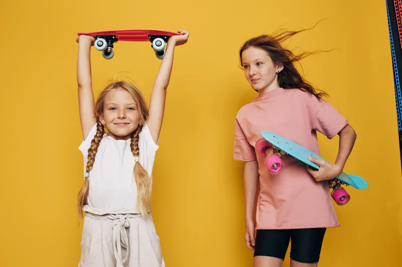Two girls with skateboards pose against a bright yellow background. One holds skateboard overhead, the other holds it sideways. Concept for youth and sports.