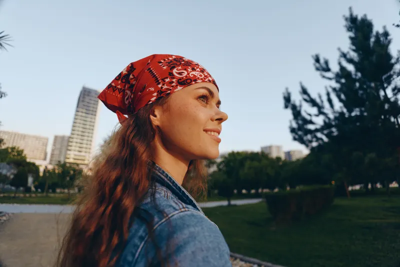 Lifestyle woman in modern boho-western denim street style wearing a red bandana smiling outdoors with film color effect and urban background during golden hour.
